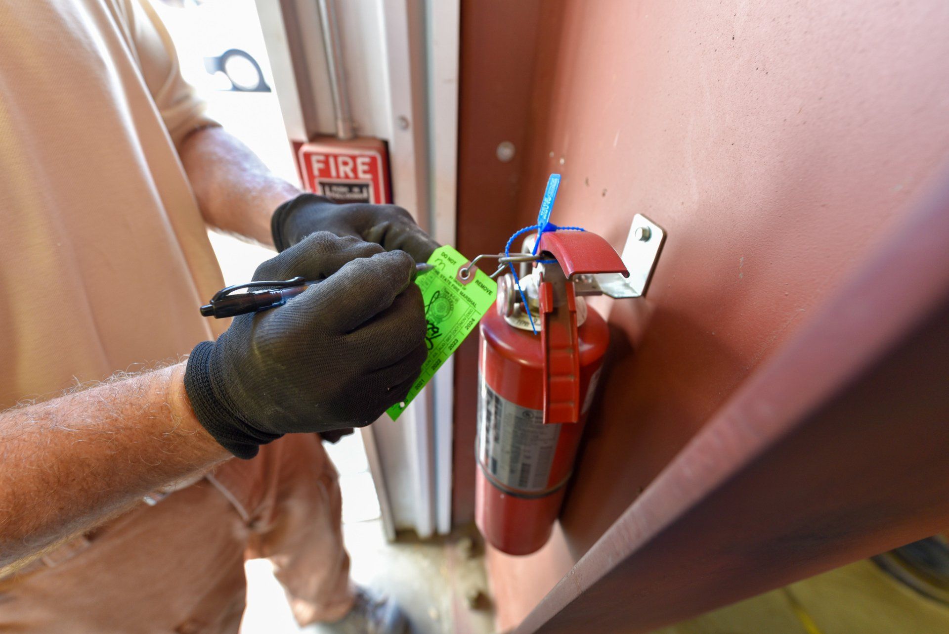 Person securing a green tamper seal on a red fire extinguisher mounted on a wall