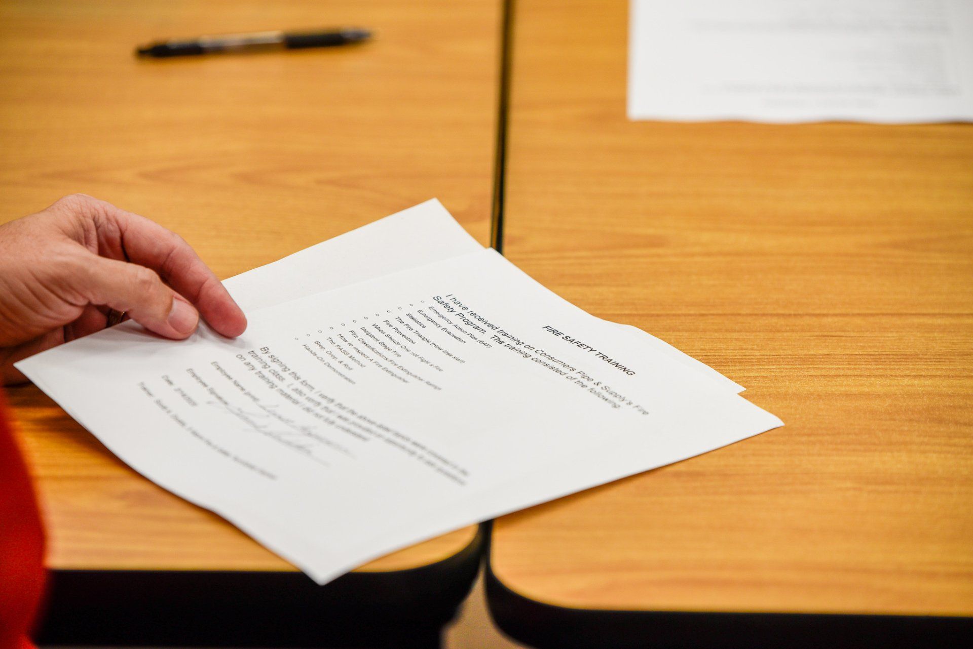 Hand holding a document over a wooden desk with papers and a pen in the background