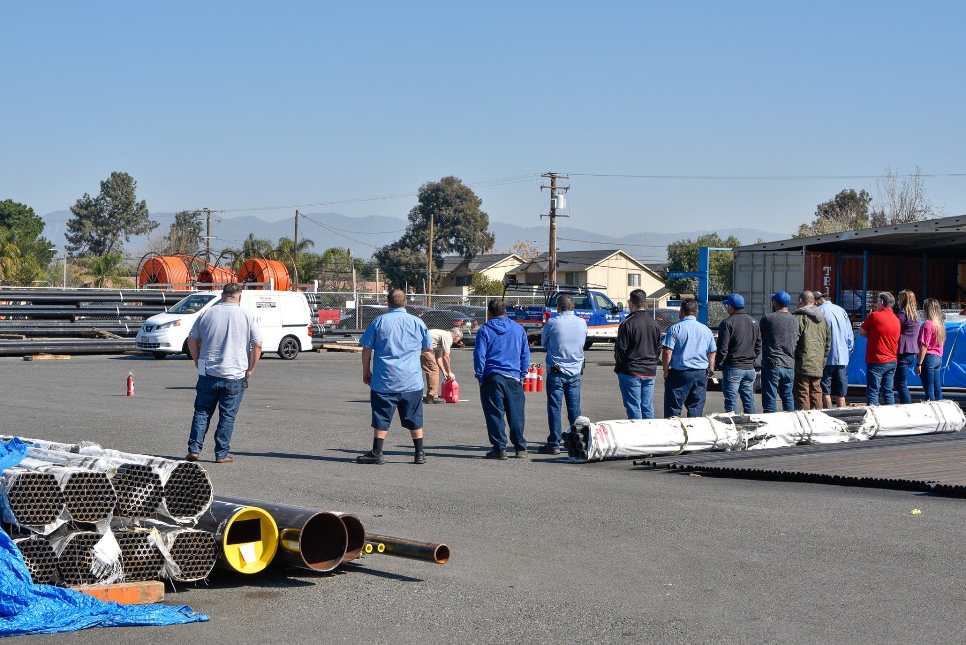 Group of people in blue shirts gathered on a paved lot beside white barriers and equipment under a clear sky