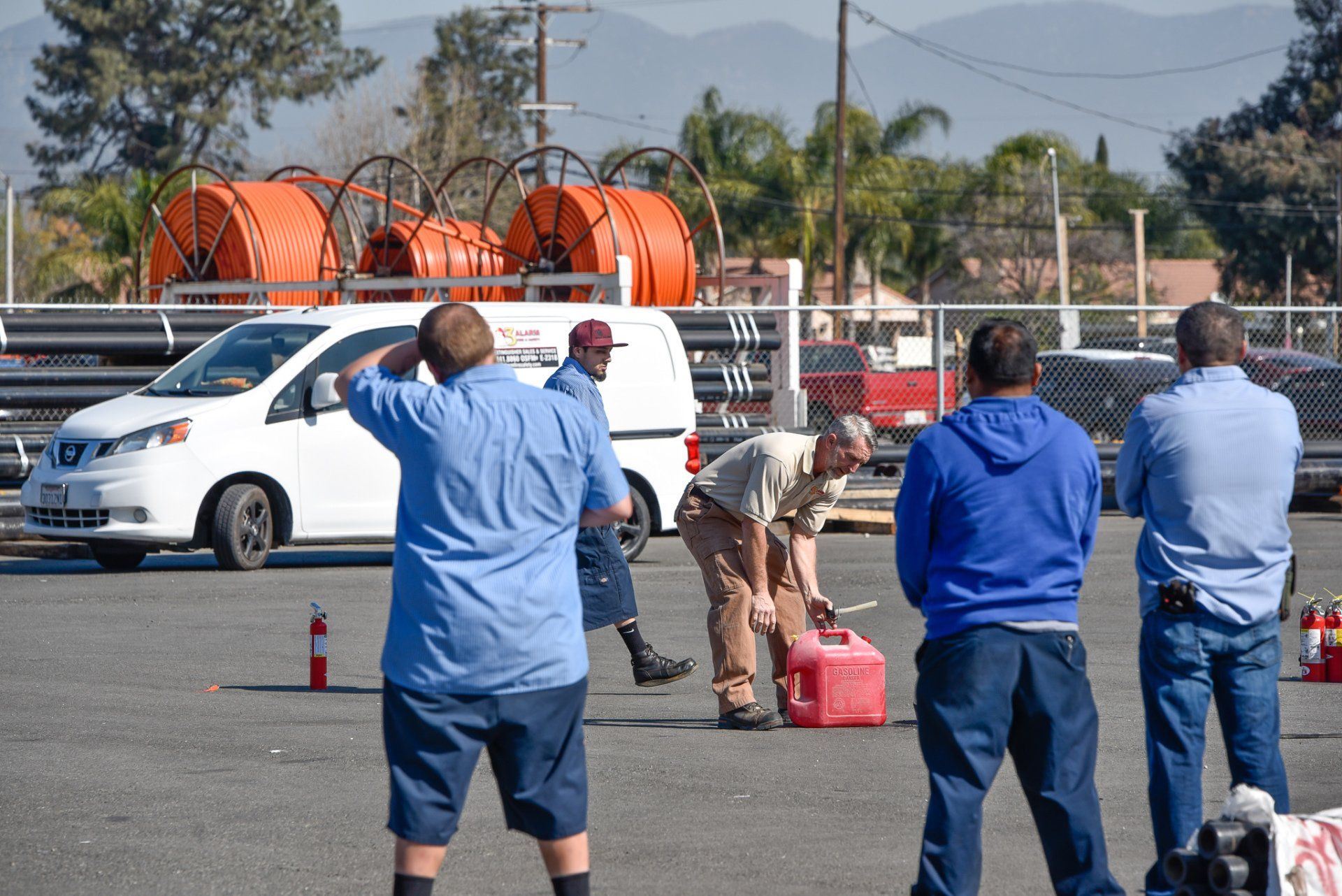Workers and bystanders near a white van at a roadside incident, with cones and red fuel cans nearby.