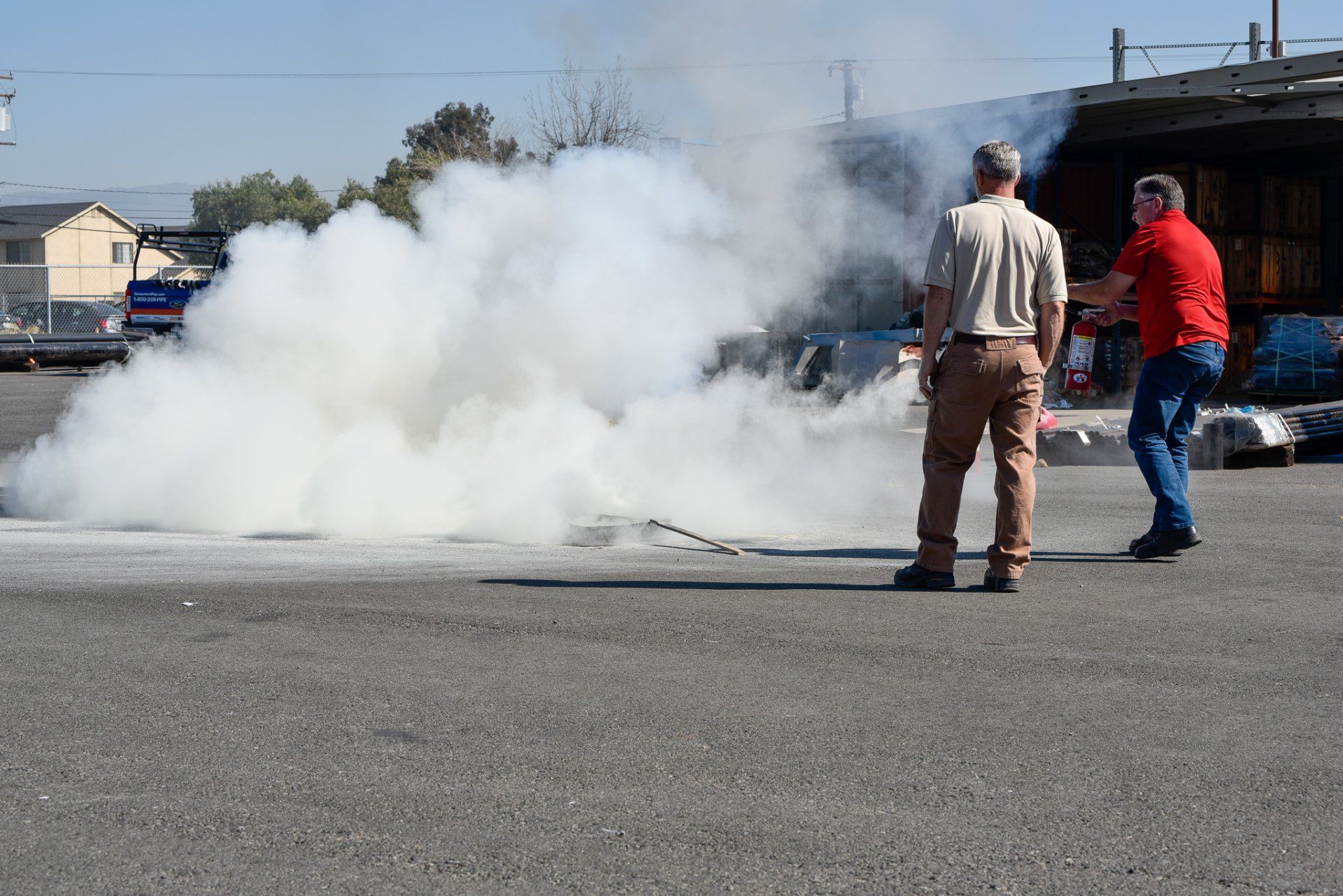 Two people stand near a large cloud of white smoke rising from the pavement in a parking lot.