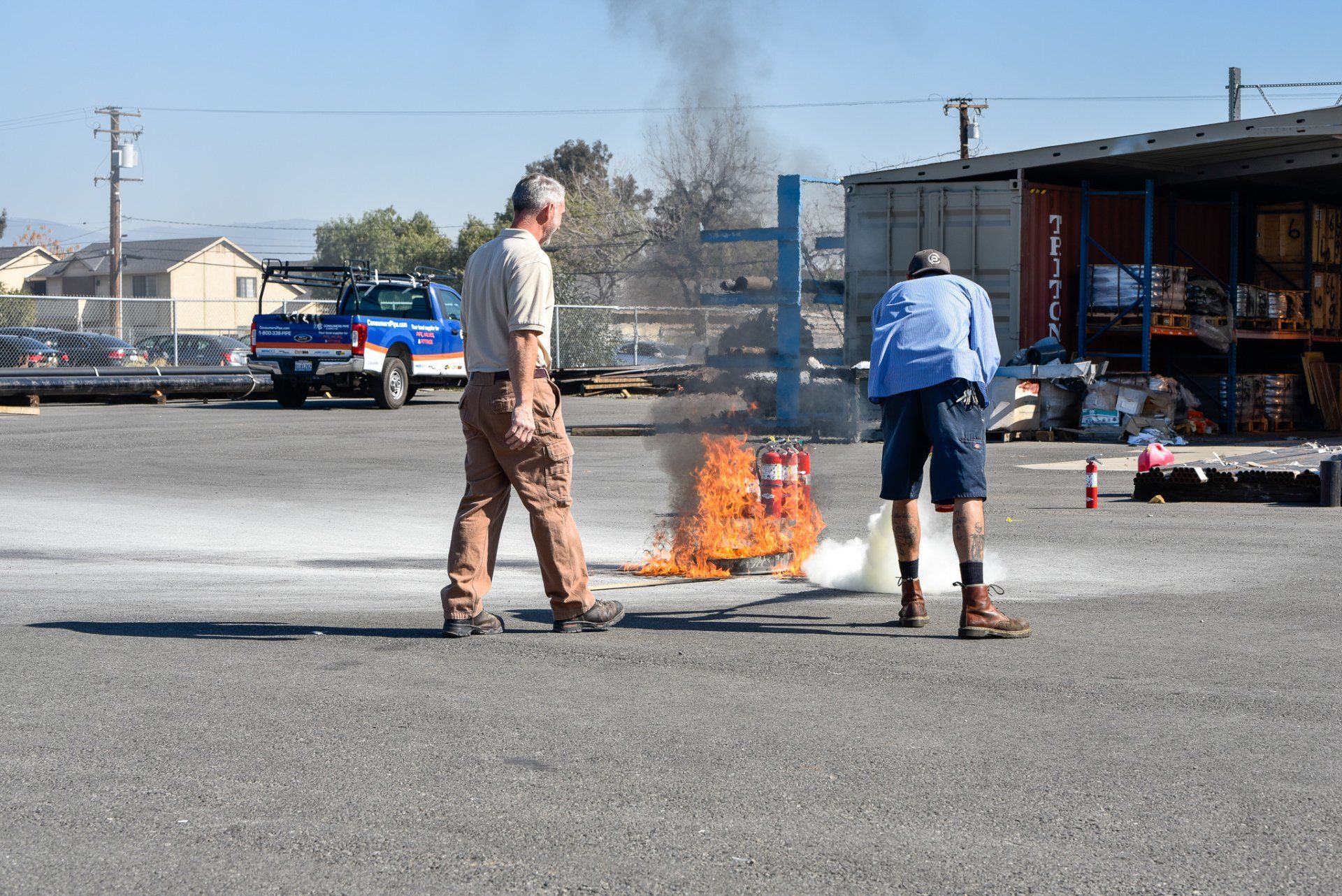 Two people stand near a small fire burning in a parking lot beside a building.