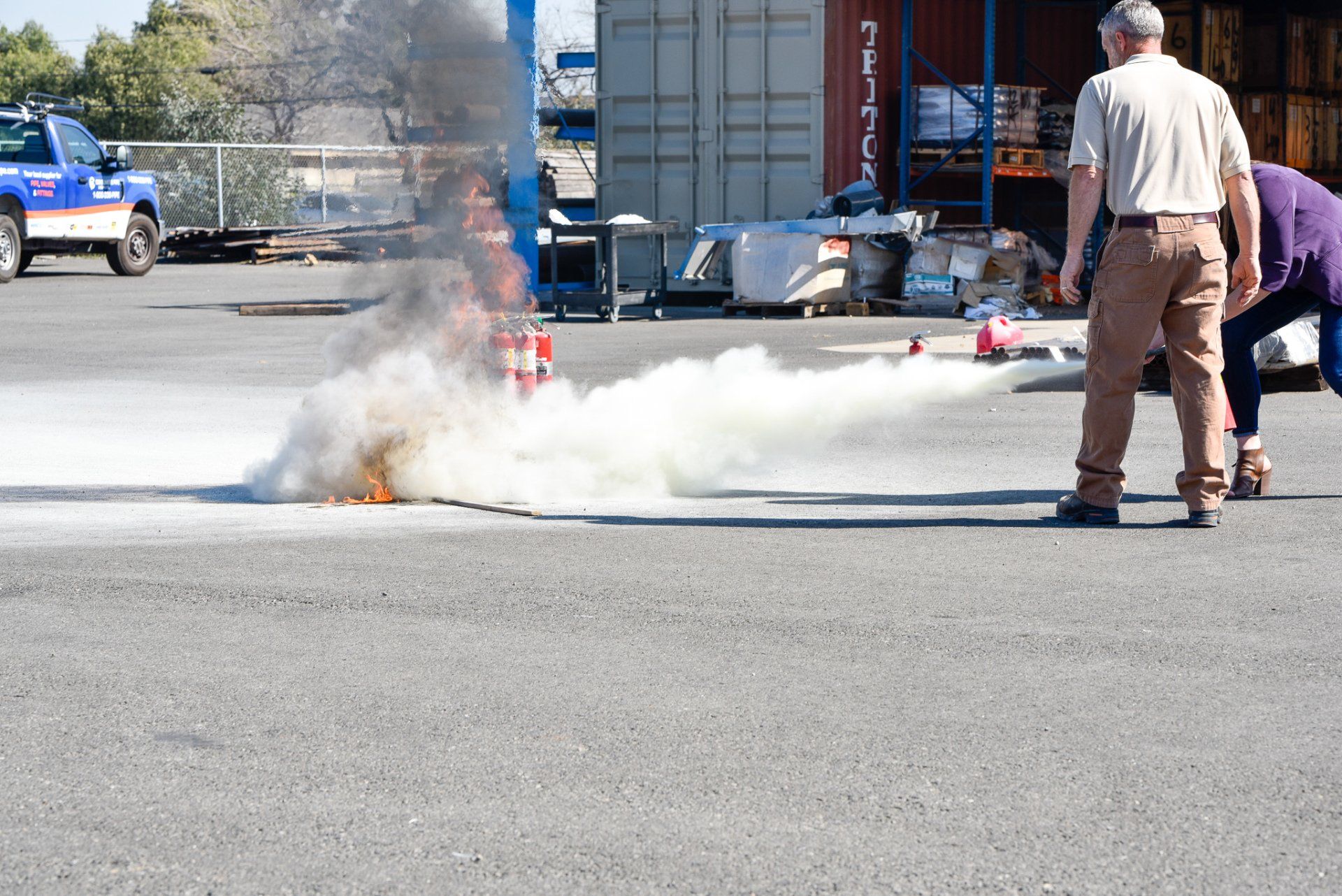 Fire and thick white smoke on a street as a man stands nearby with a fire extinguisher.