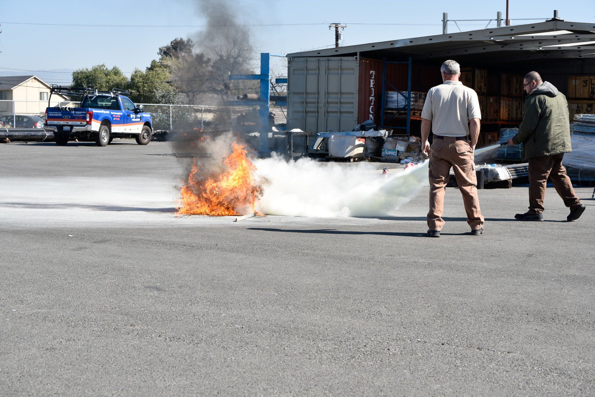 Two firefighters near a small car fire in a parking lot, with smoke rising beside a building.