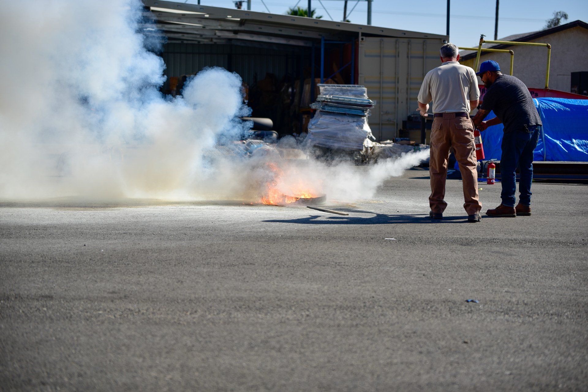 Two workers near a small ground fire with thick white smoke outside an industrial building