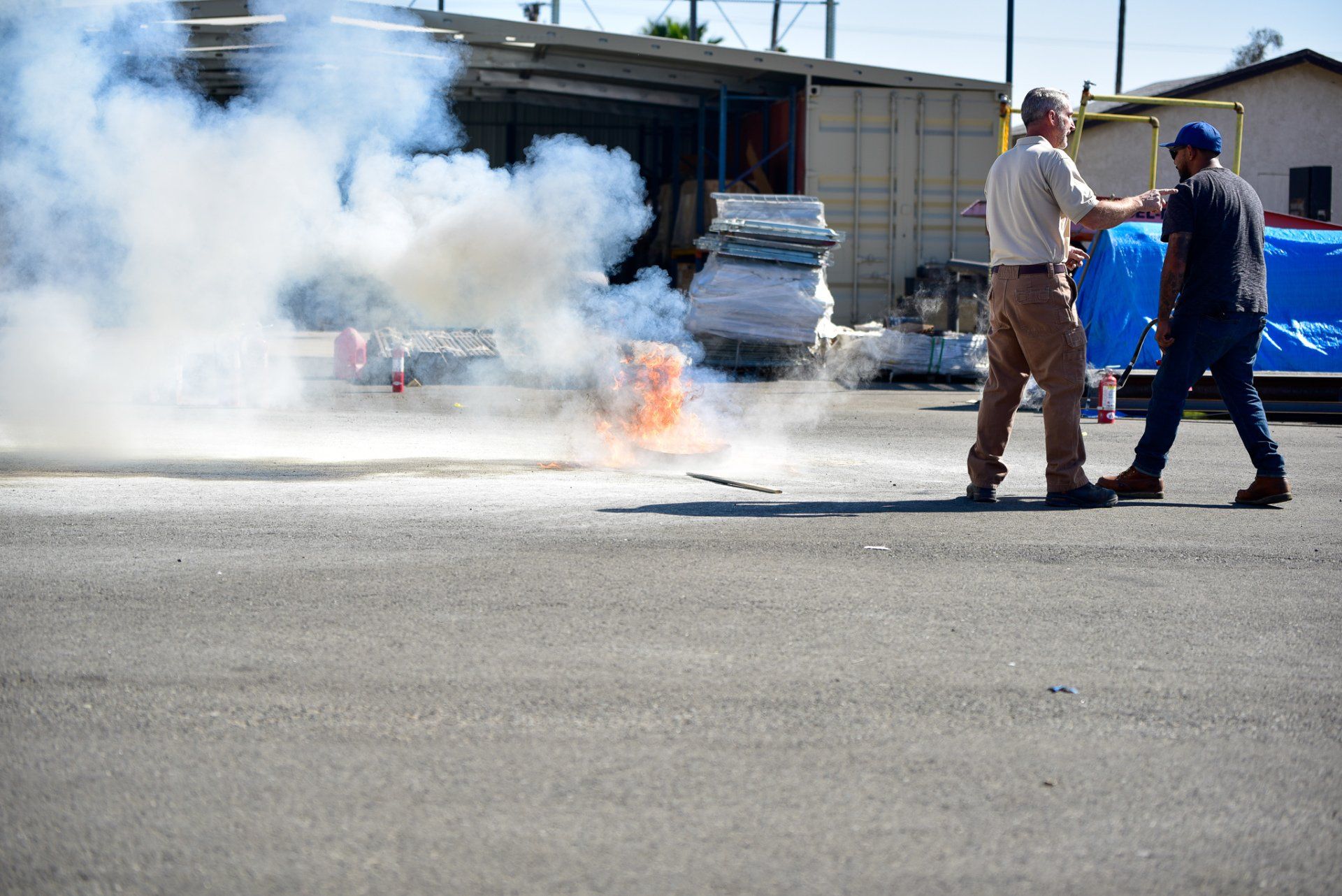 Two workers near a smoking car fire on a road, with flames and thick white smoke.