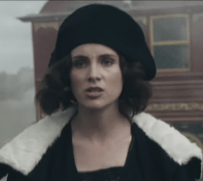 A person with dark wavy hair wears a black hat and a black coat with a white fur collar, appearing serious and focused.