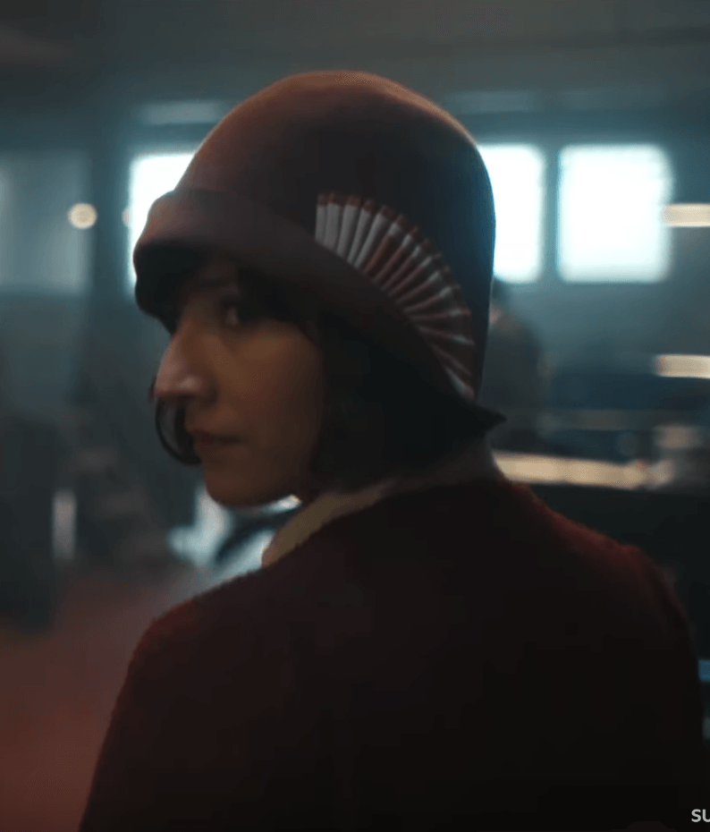 A person in a 1920s-style felt cloche hat with a decorative, fan-like trim glances over their shoulder in a dimly lit room.