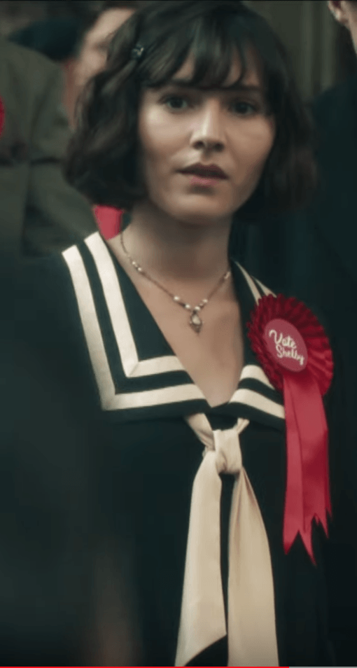 A person with short, dark hair wearing a black dress with a white sailor collar and a red rosette pinned to their chest.