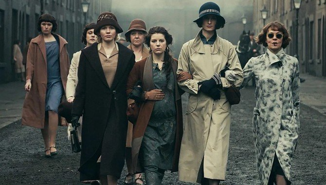 A group of people wearing 1920s-style coats and hats walk together down a cobblestone street in a somber, muted setting.