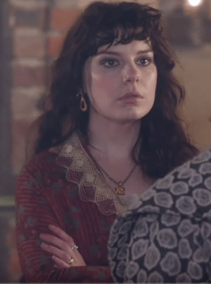 A person with dark, wavy hair and bangs wears a patterned red garment with a lace collar and earrings, arms crossed.