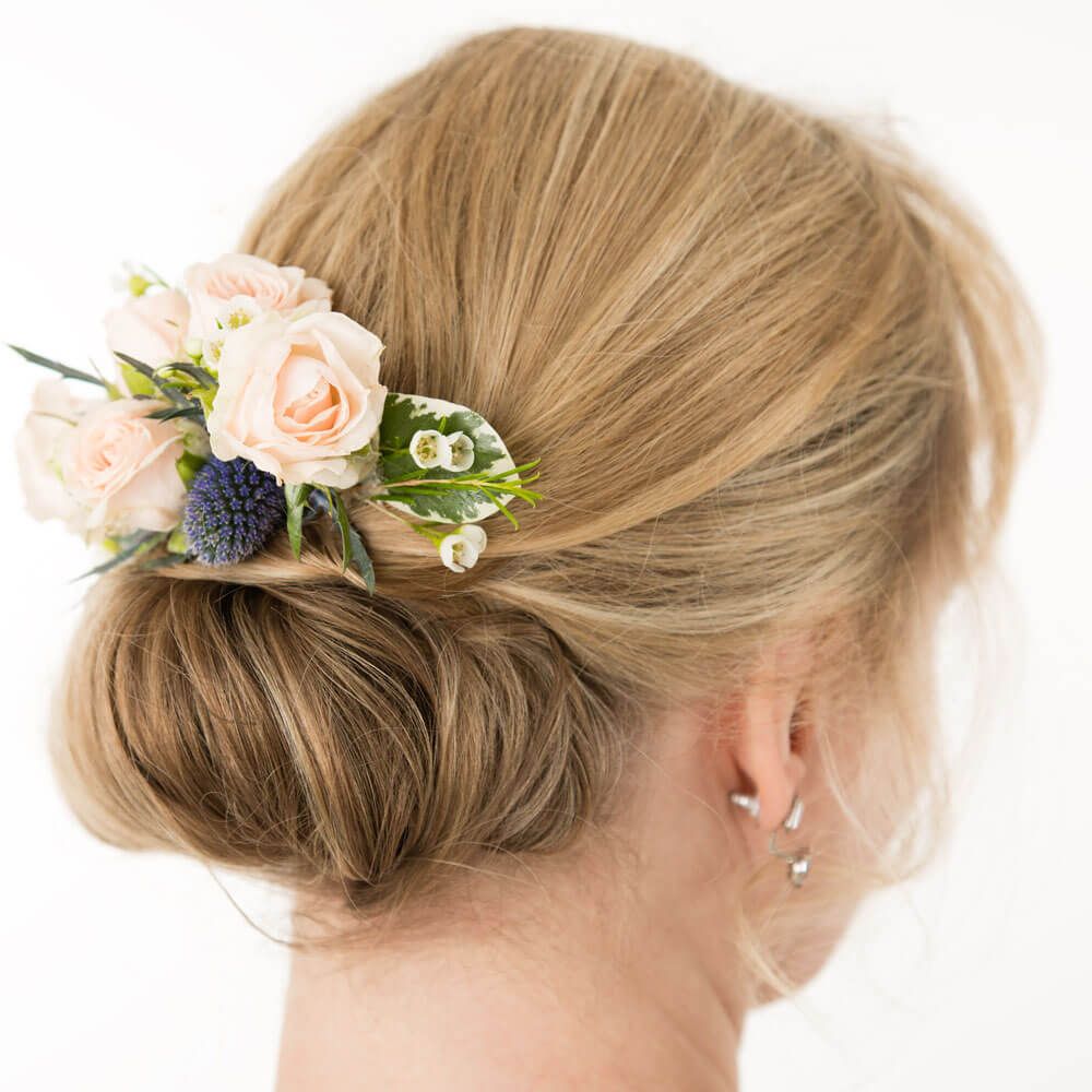 A low bun hairstyle adorned with small light pink roses, a blue thistle, and green leaves on the side.