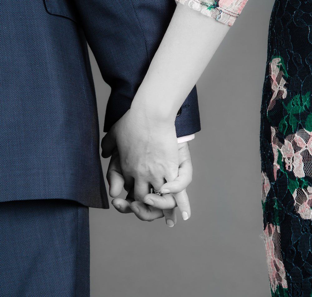 A close-up of two people holding hands, one wearing a dark blue suit and the other wearing a floral print garment.