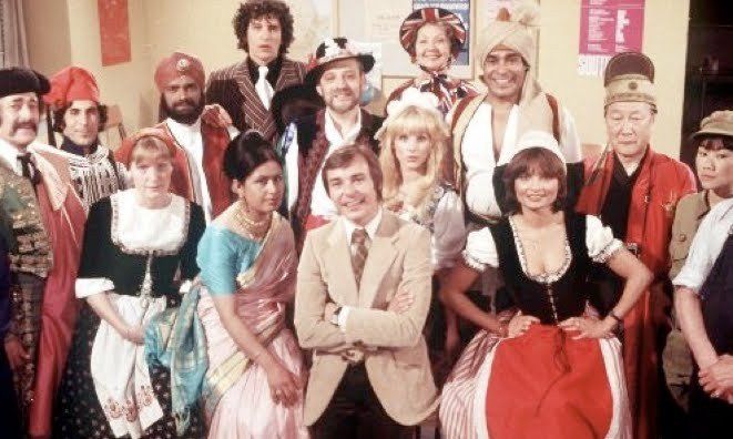 A group of people in varied cultural costumes stands around a man in a tan blazer in a room with a bulletin board.