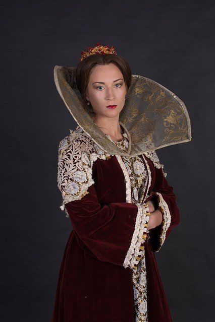 A person in a historical-style velvet gown with a large, upright lace collar and crown, posing against a dark background.