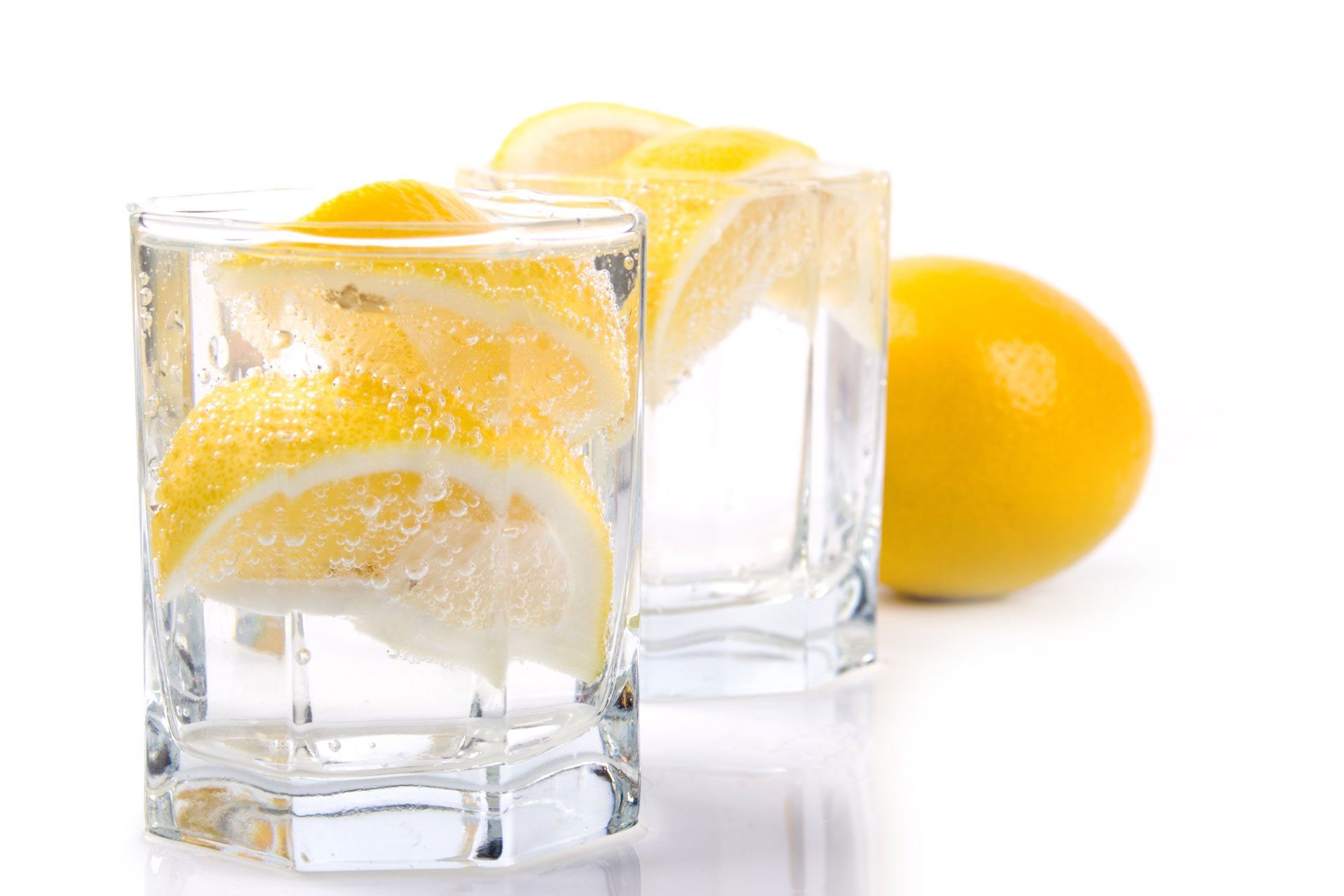 Two clear glasses filled with water and lemon wedges, with a whole lemon in the background against a white backdrop.