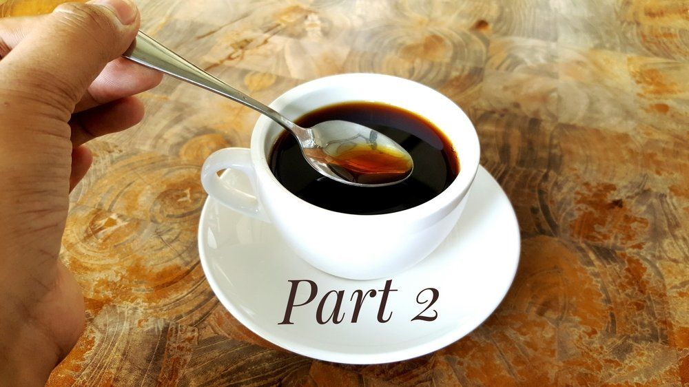 A hand holds a silver spoon over a white cup of black coffee on a saucer with