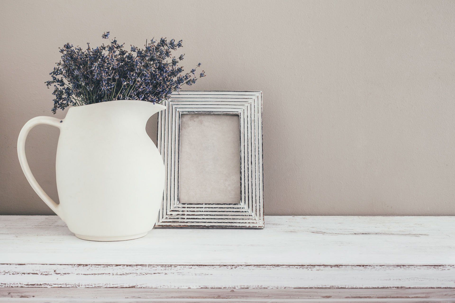 A white pitcher filled with dried lavender sits next to a rustic, white-washed empty picture frame on a wooden surface.