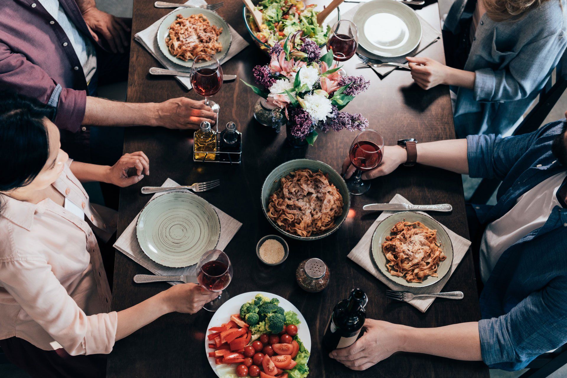Overhead view of four people sitting around a dark wooden table sharing a meal of pasta, salad, and red wine.