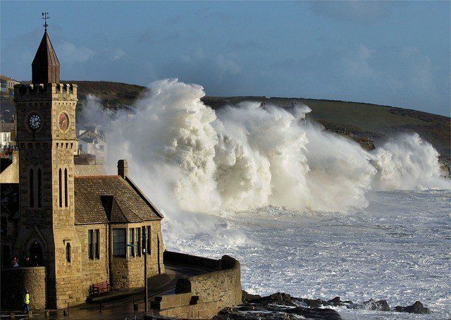 A stone building with a clock tower stands on a coastline as a massive ocean wave crashes against the shore nearby.