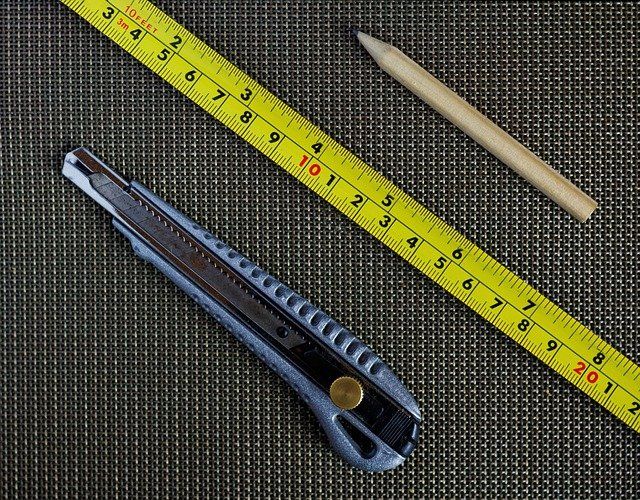A yellow measuring tape, a utility knife, and a wooden pencil arranged on a dark, textured surface.