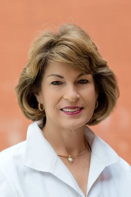 Sallie Deen wearing a white shirt and a necklace is smiling for the camera.