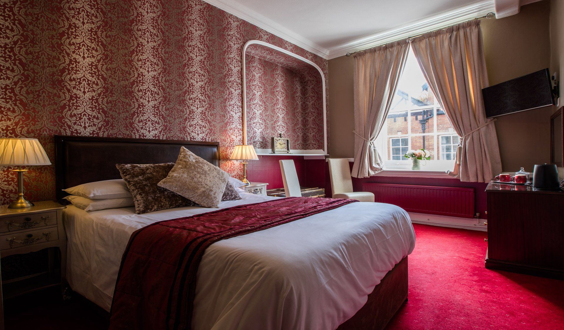 Hotel bedroom with a red patterned wallpaper and carpet, a bed, and a window.