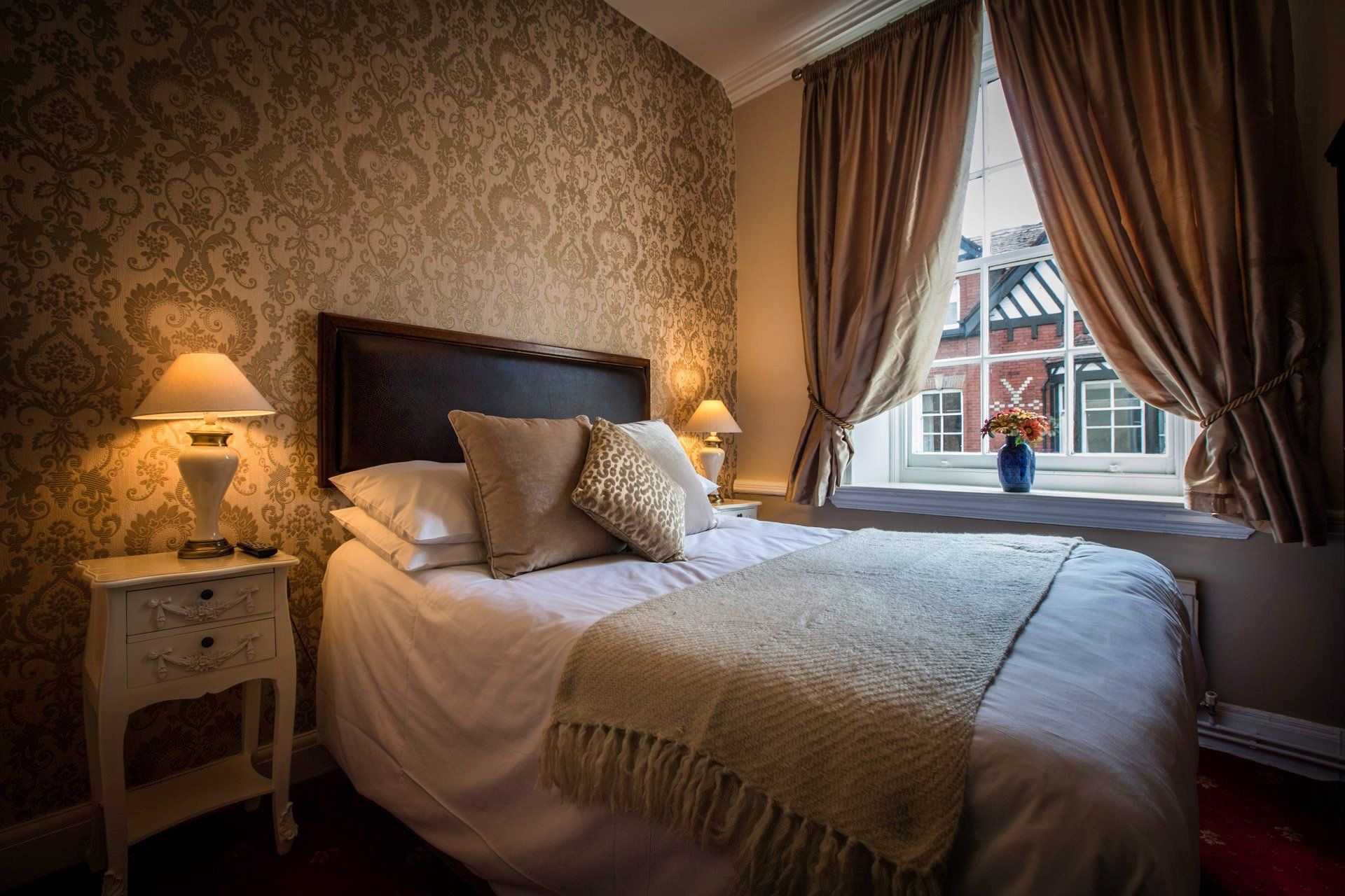 Bedroom with double bed, nightstand, patterned wallpaper, window with curtains, and two lamps.