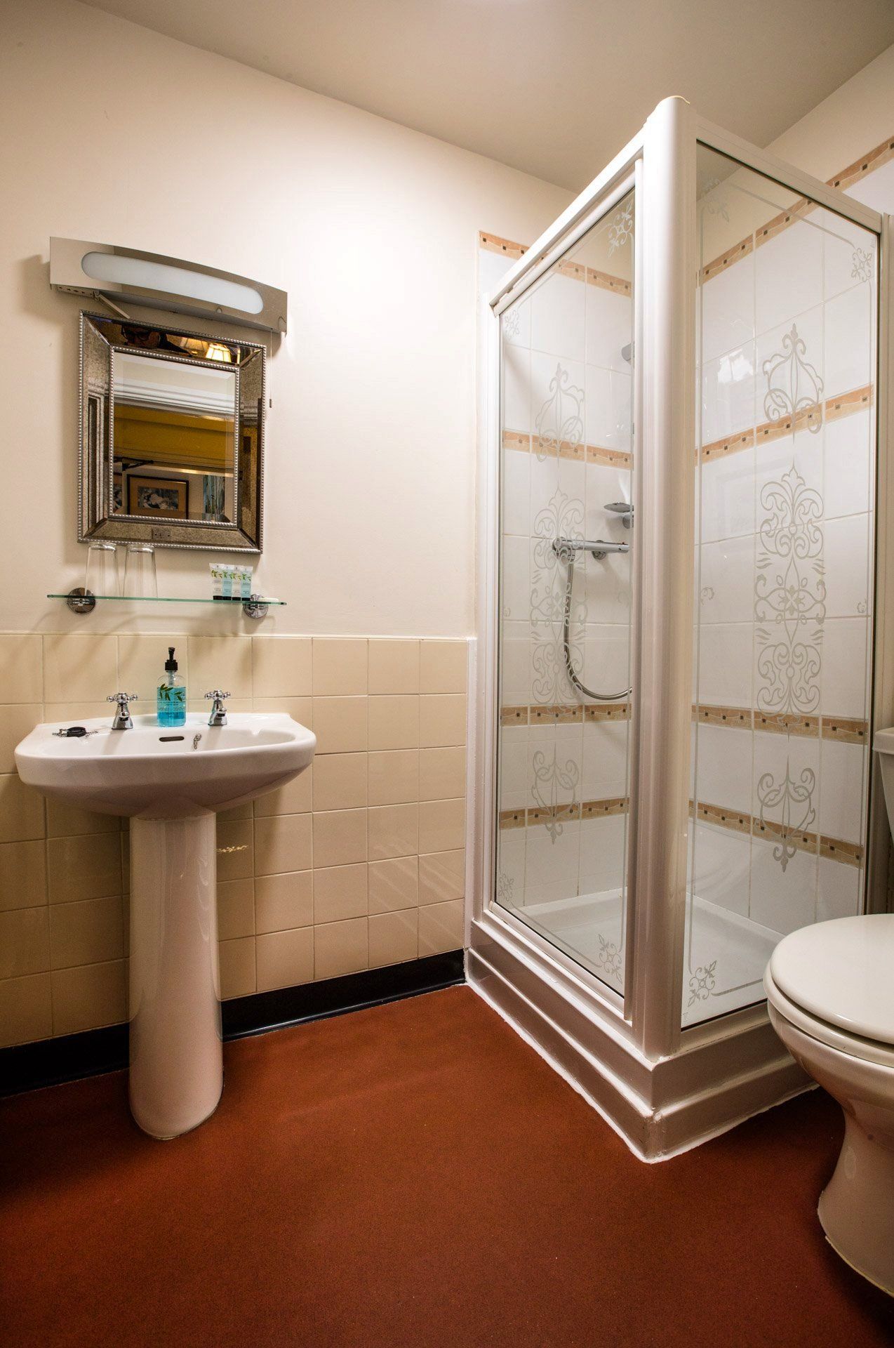 Bathroom with a pedestal sink, shower with glass door, mirror, and toilet. Red floor, beige tiles.