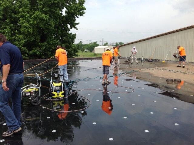 Workers in orange vests pressure-wash a wet rooftop beside a beige building.