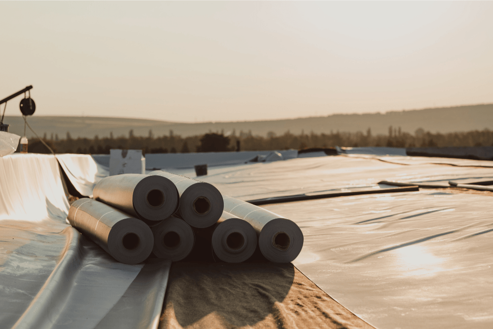 Rolls of black roofing membrane laid out on a sunlit flat roof with plastic sheeting nearby
