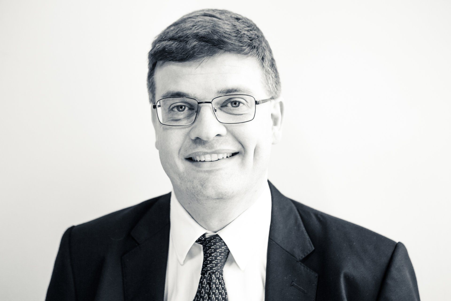 Professional headshot of a a gentelman wearing glasses and suit, stood against a light grey background