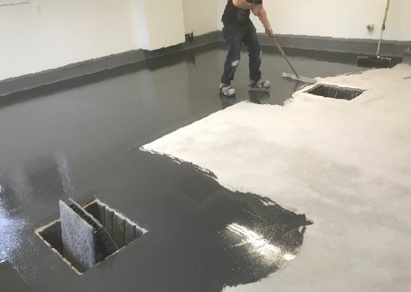 Person applying dark epoxy floor coating. White wall, two square openings, unfinished area.