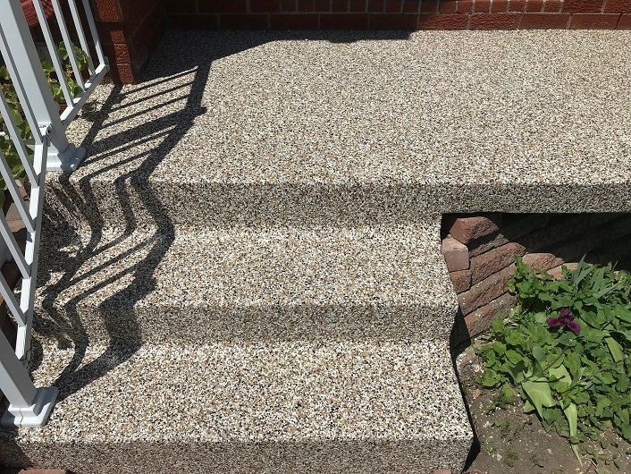 Stone-textured steps leading up to a front porch. White railing on the left, garden on the right.