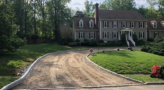 Driveway being worked on in front of a two-story house, bordered by grass and trees.
