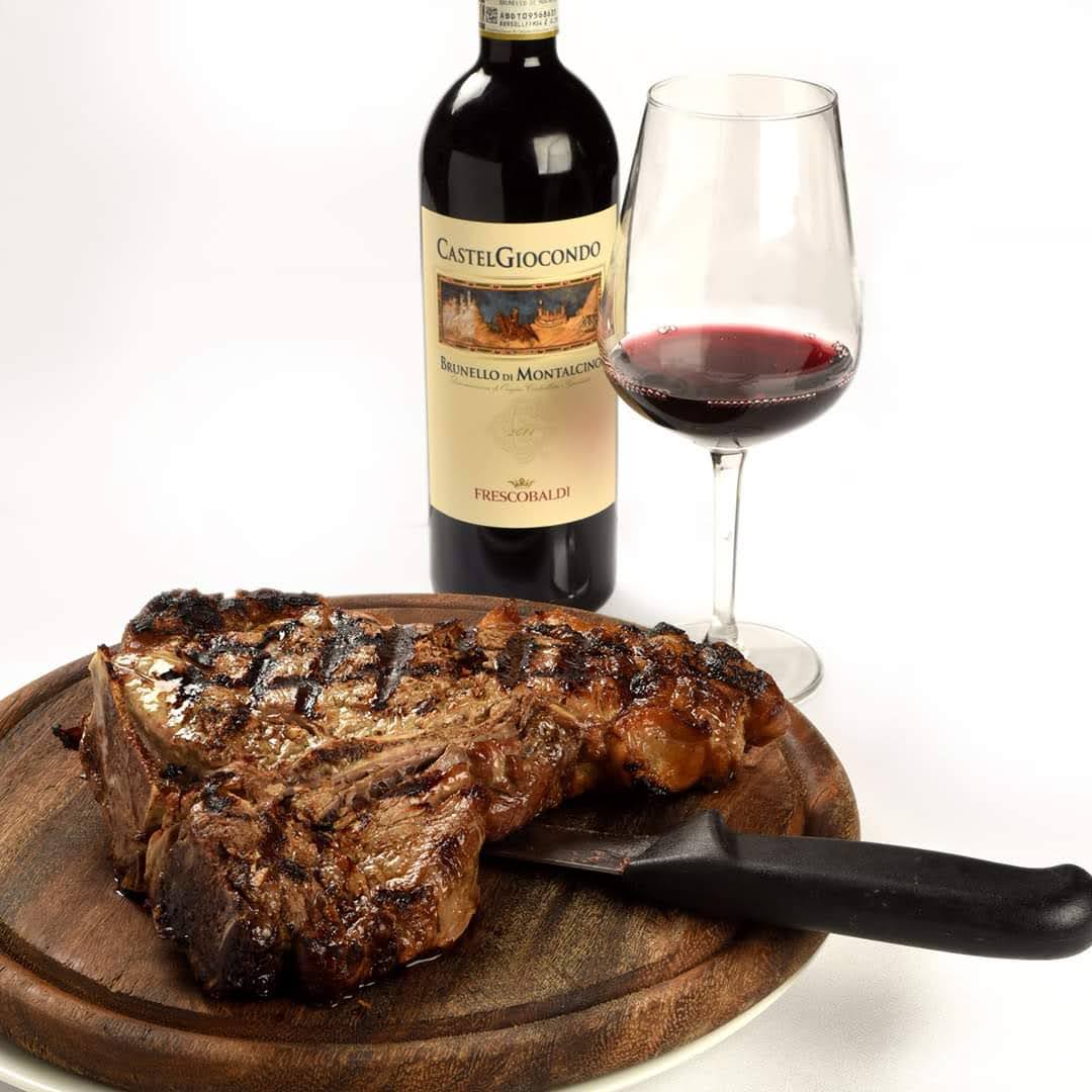 Bistecca alla fiorentina grigliata su un tagliere di legno, con una bottiglia di vino e un bicchiere di vino rosso.
