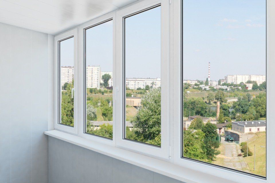 Balcone con finestre con cornice bianca che si affacciano su un paesaggio urbano con alberi verdi ed edifici sotto un cielo azzurro.