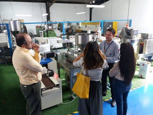 Personas recorriendo una planta de producción con máquinas y un guía en un luminoso espacio de trabajo industrial.