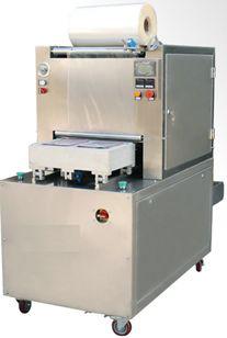 Máquina industrial de acero inoxidable para retractilado, con rollo de film en la parte superior y bandeja transportadora sobre ruedas.