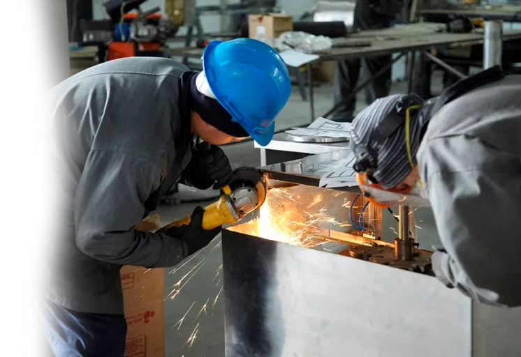 Trabajadores con cascos de seguridad soldando metal en un taller industrial; saltan chispas.
