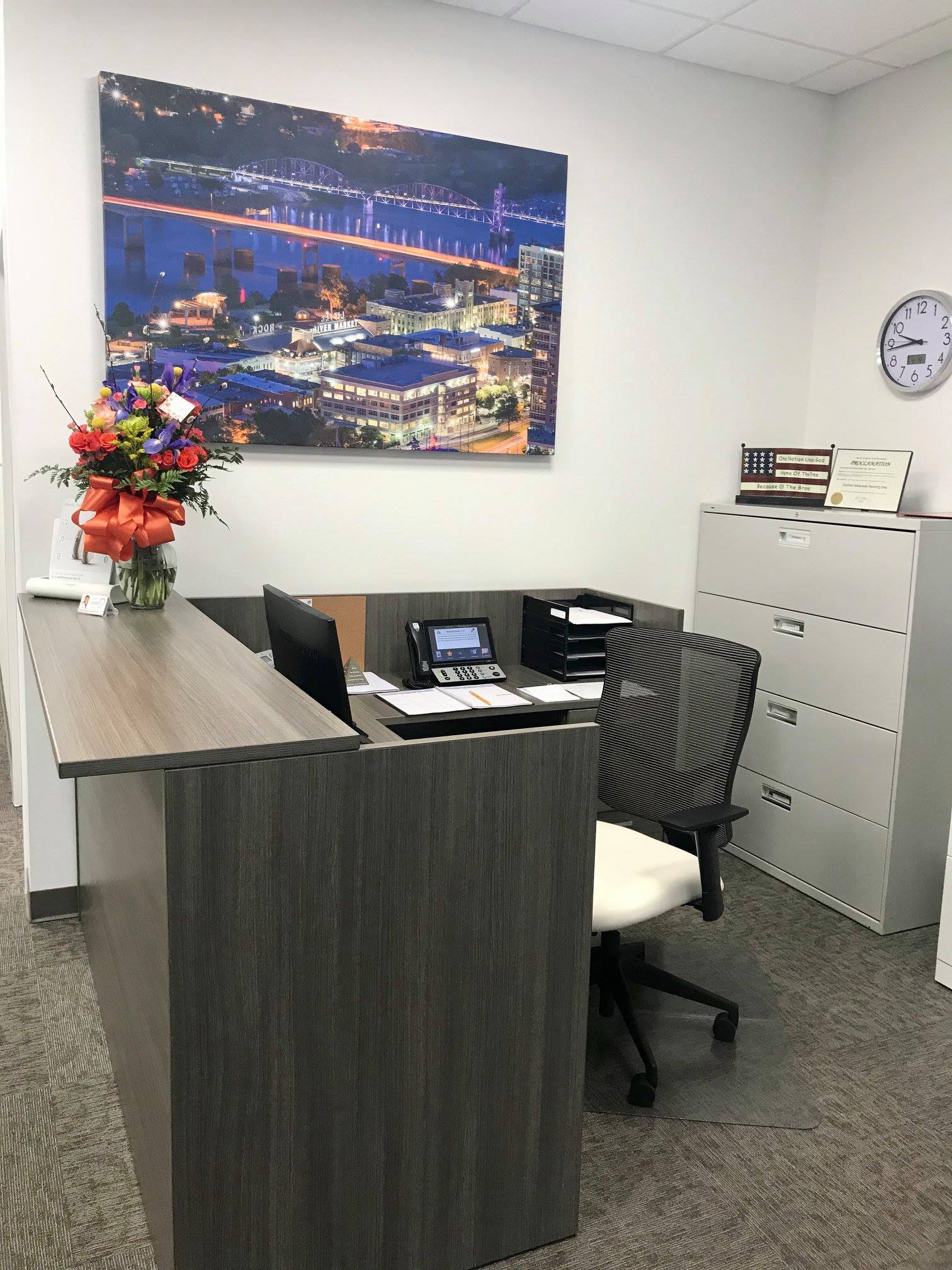 Small office reception desk with chair, filing cabinet, wall art, and flowers.