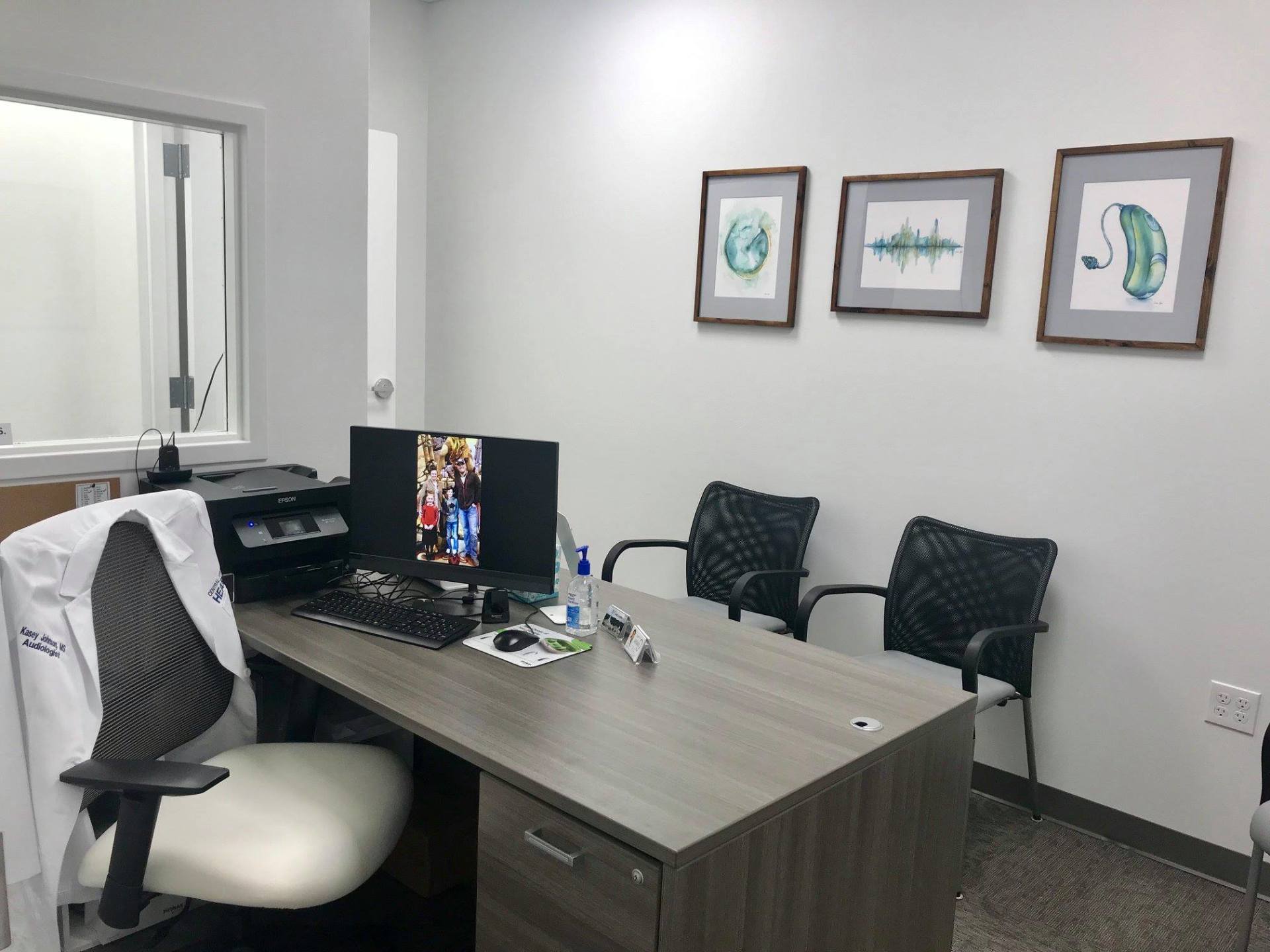 Office desk with computer, chairs, and framed artwork on a white wall
