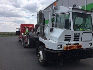 White yard truck towing a red tow truck outdoors on a paved surface. The setting appears to be an industrial area.