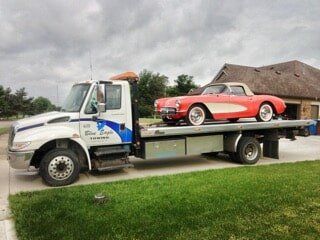 A red vintage Corvette is being towed on the flatbed of a white tow truck, parked on a driveway in front of a house.