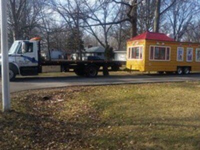 A yellow tiny house with a red roof is towed by a flatbed truck on a street with a grassy yard.