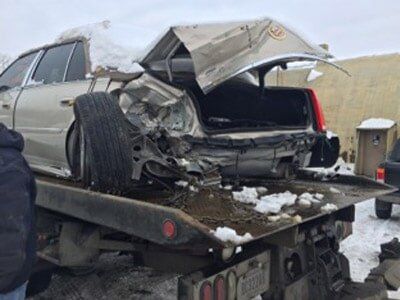 A severely damaged silver sedan being towed after a collision. Its trunk is crumpled, and the rear is crushed.