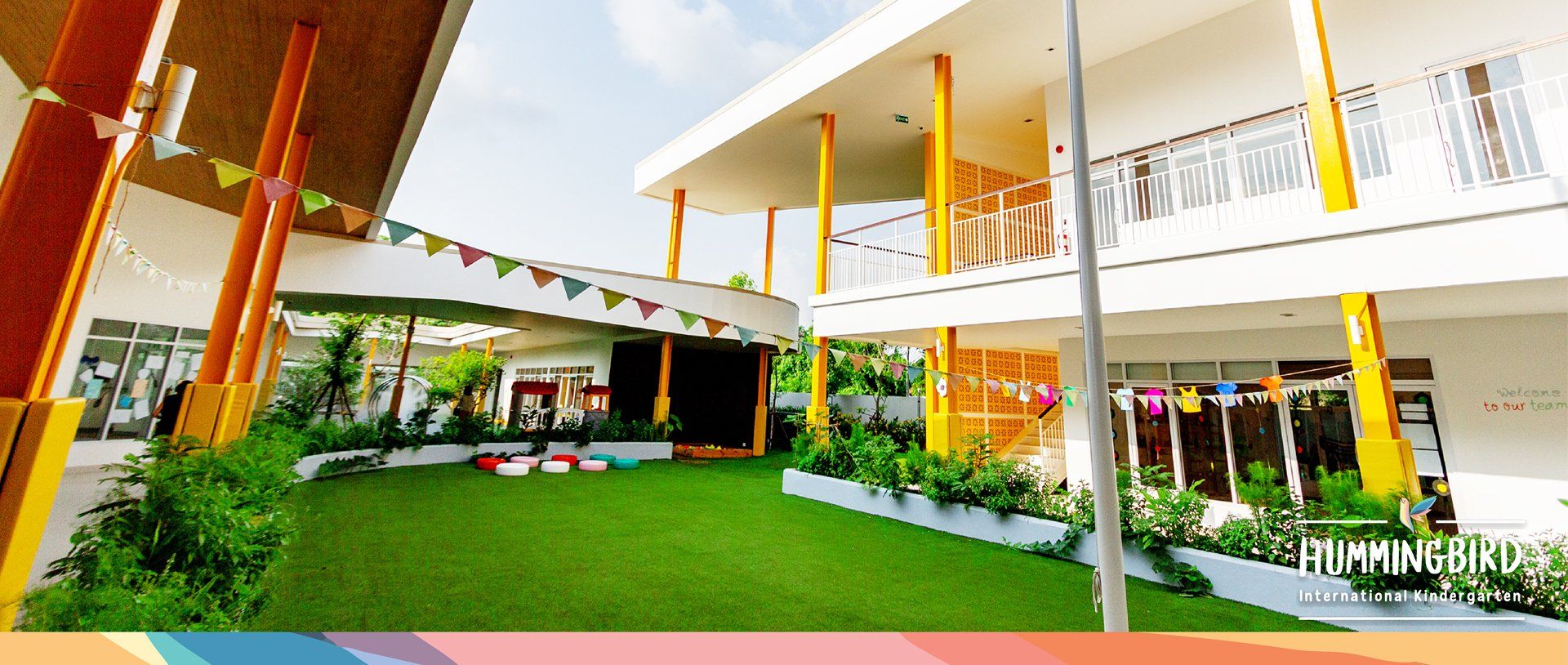 มุมมองภายนอกของอาคารทันสมัยที่สดใสพร้อมสนามหญ้าสีเขียว ธงสีสันสดใส และเสาสีเหลือง