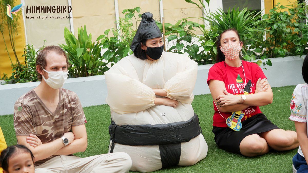 A group of people wearing face masks are sitting on the grass next to a sumo wrestler.