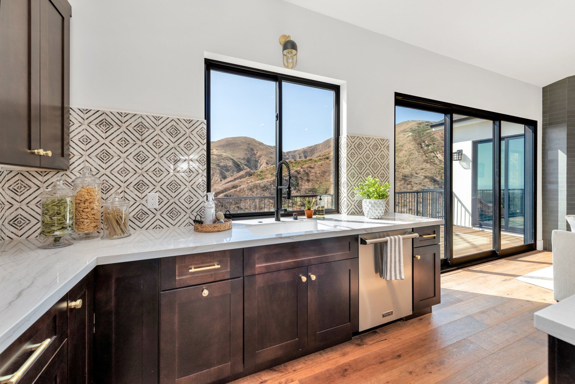 An open kitchen with a sink, dishwasher, sliding glass doors, dark colored cabinets, and white marble countertops.