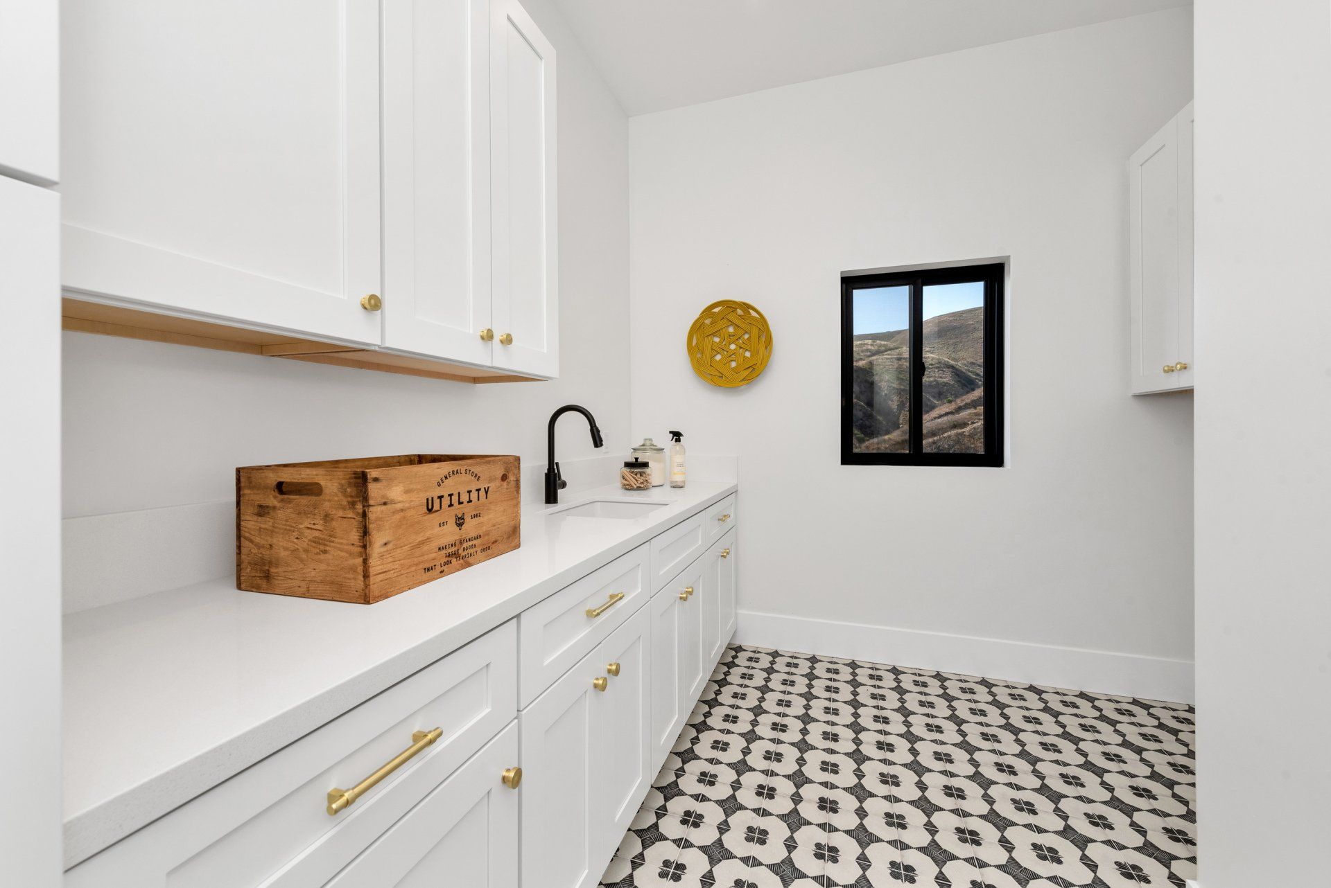 A laundry room with white cabinets, white counters, a sink, and a window with a wooden box on the counter.